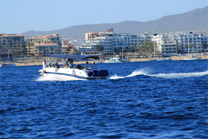 Motorboat with a canopy and passengers cutting through deep blue water near a sandy beach and marina, framed by beachfront resort buildings and distant hills on a sunny coastal getaway.