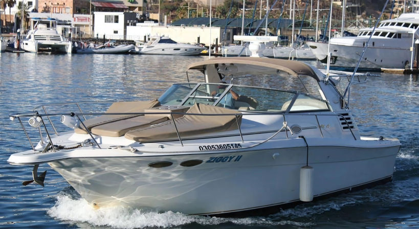 Sleek white cabin cruiser with tan sunpads and bimini gliding through blue harbor water past a busy marina with moored yachts