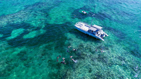 Aerial view of a white catamaran anchored over crystal-clear turquoise water and shallow coral reef with a group of snorkelers and a small dinghy nearby, tropical ocean scene.