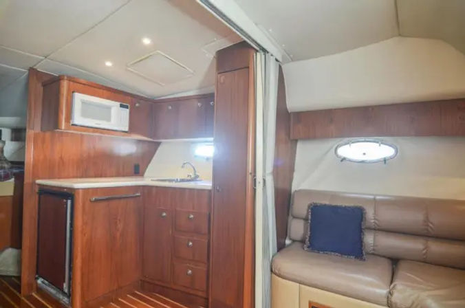 Cozy yacht cabin galley interior with warm wood cabinetry, compact sink and microwave, mini fridge, tan leather sofa with navy throw pillow and oval porthole window.