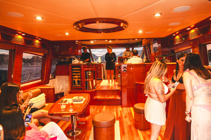 Luxury wood-paneled yacht cabin with bar and seating, small group socializing and sipping drinks, large windows showing a coastal harbor at dusk