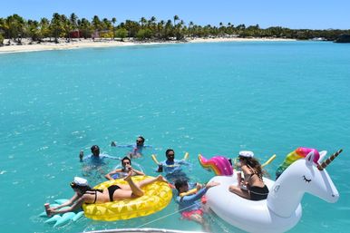 Group of people lounging and splashing on inflatable pineapple and unicorn floats in crystal‑clear turquoise water off a palm‑lined tropical beach with colorful huts and a bright blue sky.