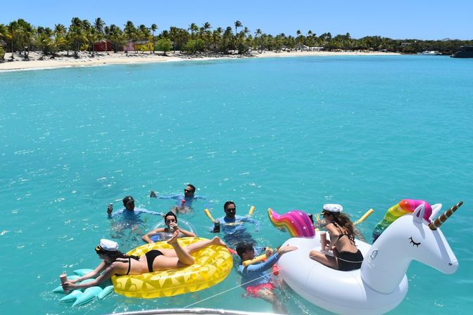 Group of people lounging and splashing on inflatable pineapple and unicorn floats in crystal‑clear turquoise water off a palm‑lined tropical beach with colorful huts and a bright blue sky.