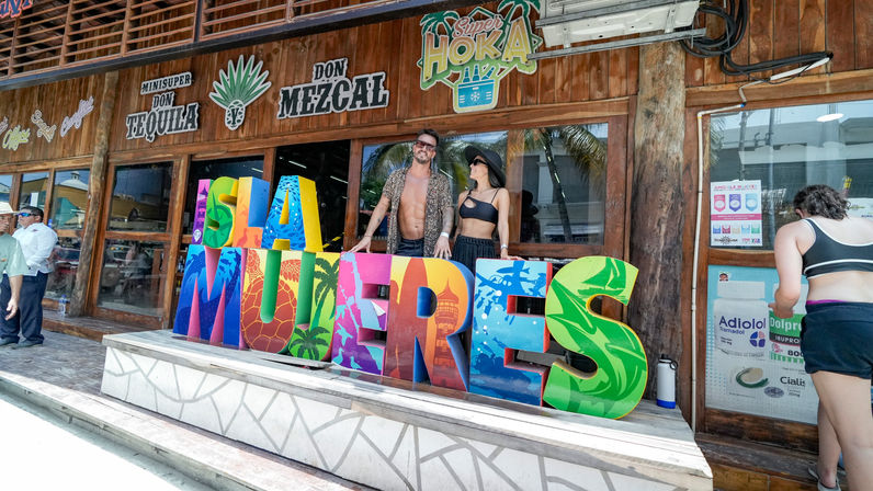 Vibrant multicolored 'Playa Mujeres' letters outside a wooden beachfront storefront in Playa Mujeres, Mexico, with two tourists in summer outfits posing behind the sign and palm trees reflected in the windows.