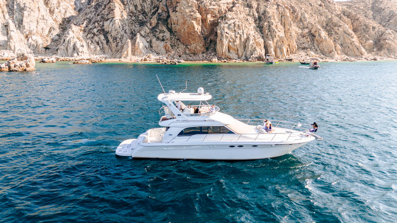 White luxury motor yacht cruising turquoise ocean near rugged rocky cliffs and a small sandy cove, people relaxing on deck on a sunny day