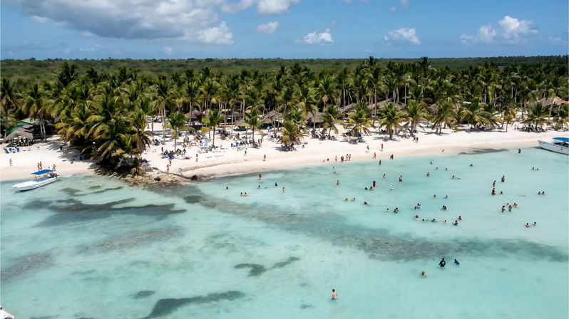 Aerial view of a tropical island palm-lined white-sand beach with turquoise shallow water, dozens of people wading and swimming and small boats on a sunny day.