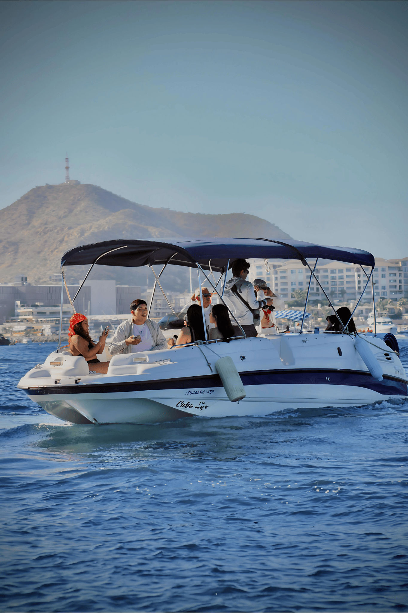 Sunny coastal boat ride: a group on a white motorboat with a navy canopy enjoying blue ocean waves, with a hillside and resort buildings in the background.