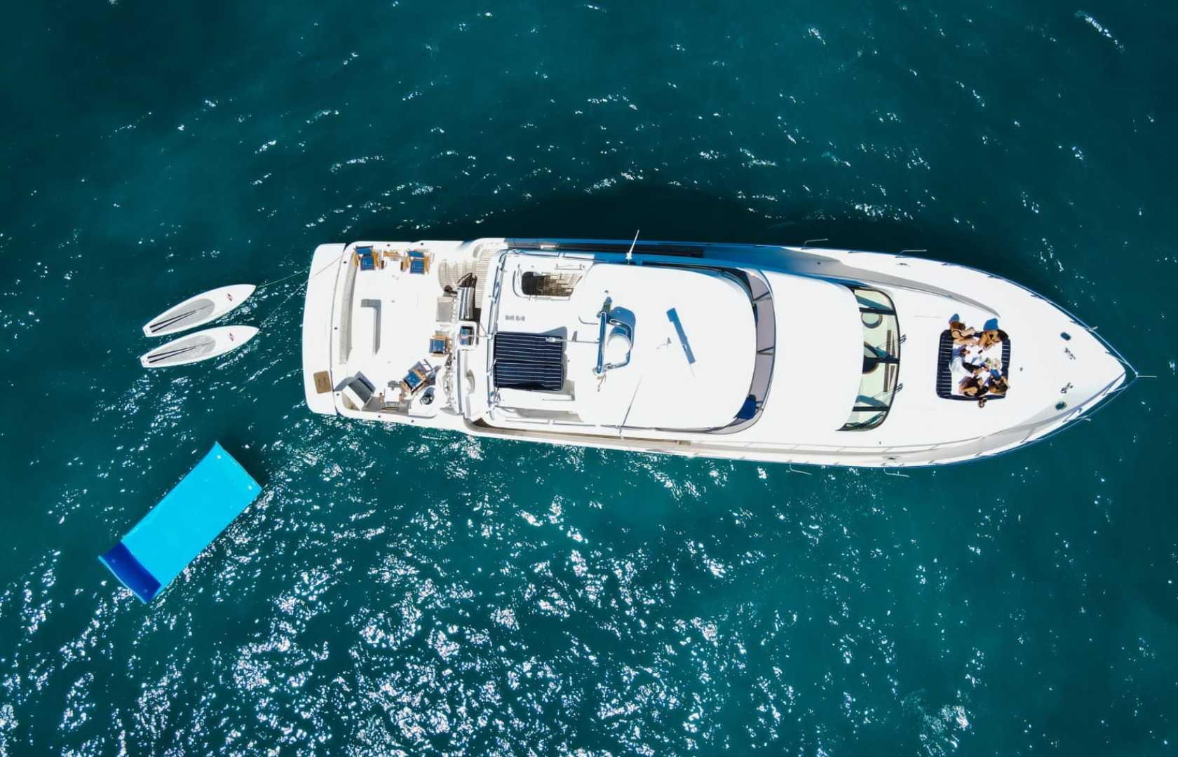 Drone view of a luxury white yacht in deep turquoise sea with a blue floating sunbed, two paddleboards, and people lounging on the bow.