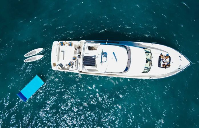 Drone view of a luxury white yacht in deep turquoise sea with a blue floating sunbed, two paddleboards, and people lounging on the bow.