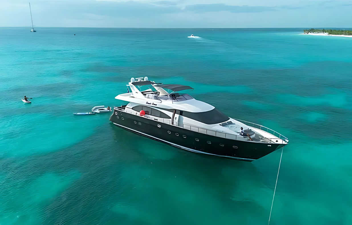 Drone view of a luxury yacht anchored in clear turquoise Caribbean waters near a small tropical island, with paddleboarders and a tender nearby.