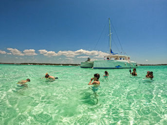 Anchored white catamaran in a shallow tropical turquoise lagoon with snorkelers wading and swimming under a bright blue sky.