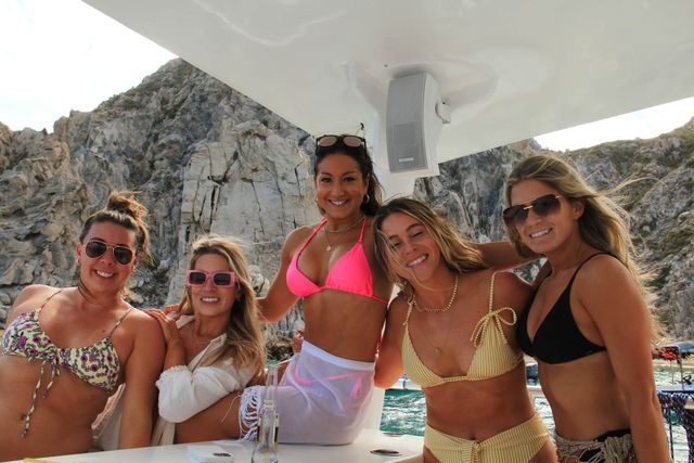 Five smiling women in colorful bikinis posing on a boat under a canopy with rocky coastal cliffs and turquoise ocean water in the background
