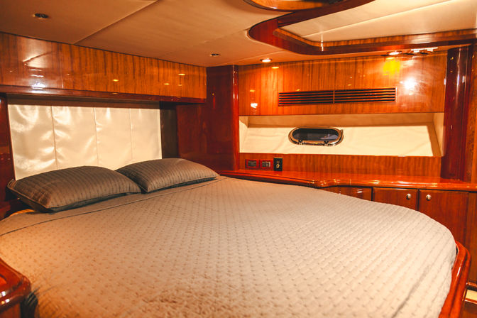 Cozy luxury yacht cabin with glossy wood paneling, queen bed, gray quilted cover and small porthole window