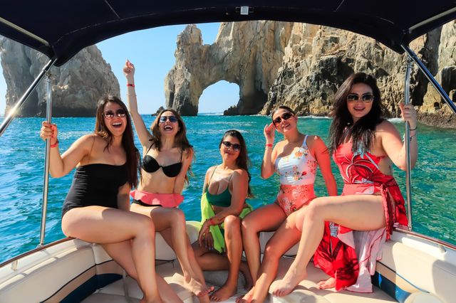 Five women in colorful swimsuits laughing on a boat with turquoise water and the iconic El Arco rock formation at Cabo San Lucas, Mexico on a bright sunny day