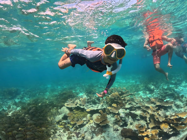 Underwater photo of snorkelers in crystal-clear tropical turquoise water above a shallow coral reef, one wearing a yellow mask and snorkel