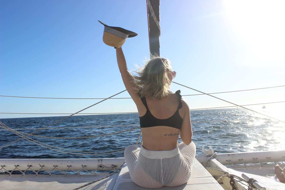 Woman sitting on the bow of a sailboat, holding a straw hat aloft with wind-tousled hair, sparkling blue ocean and bright sun under a clear sky — sunny sea vacation vibe