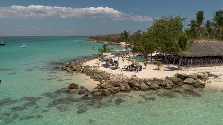 Aerial view of a tropical island beach with turquoise water, white sand and rocks, palm trees, beachfront cabanas and anchored boats under a blue sky