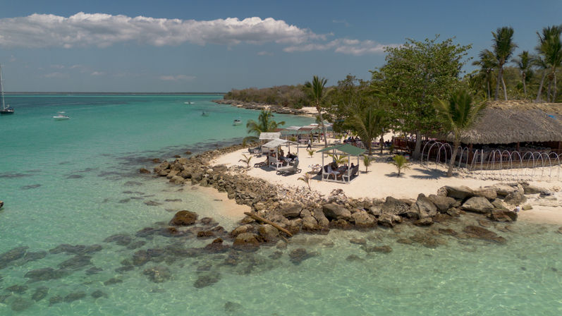 Aerial view of a tropical island beach with turquoise water, white sand and rocks, palm trees, beachfront cabanas and anchored boats under a blue sky