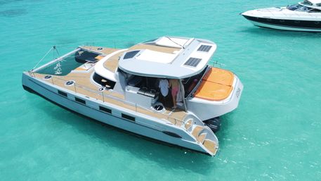 Aerial view of a white luxury catamaran yacht with teak decks and an orange sun pad, anchored in clear turquoise tropical water with two people in the shaded cockpit.