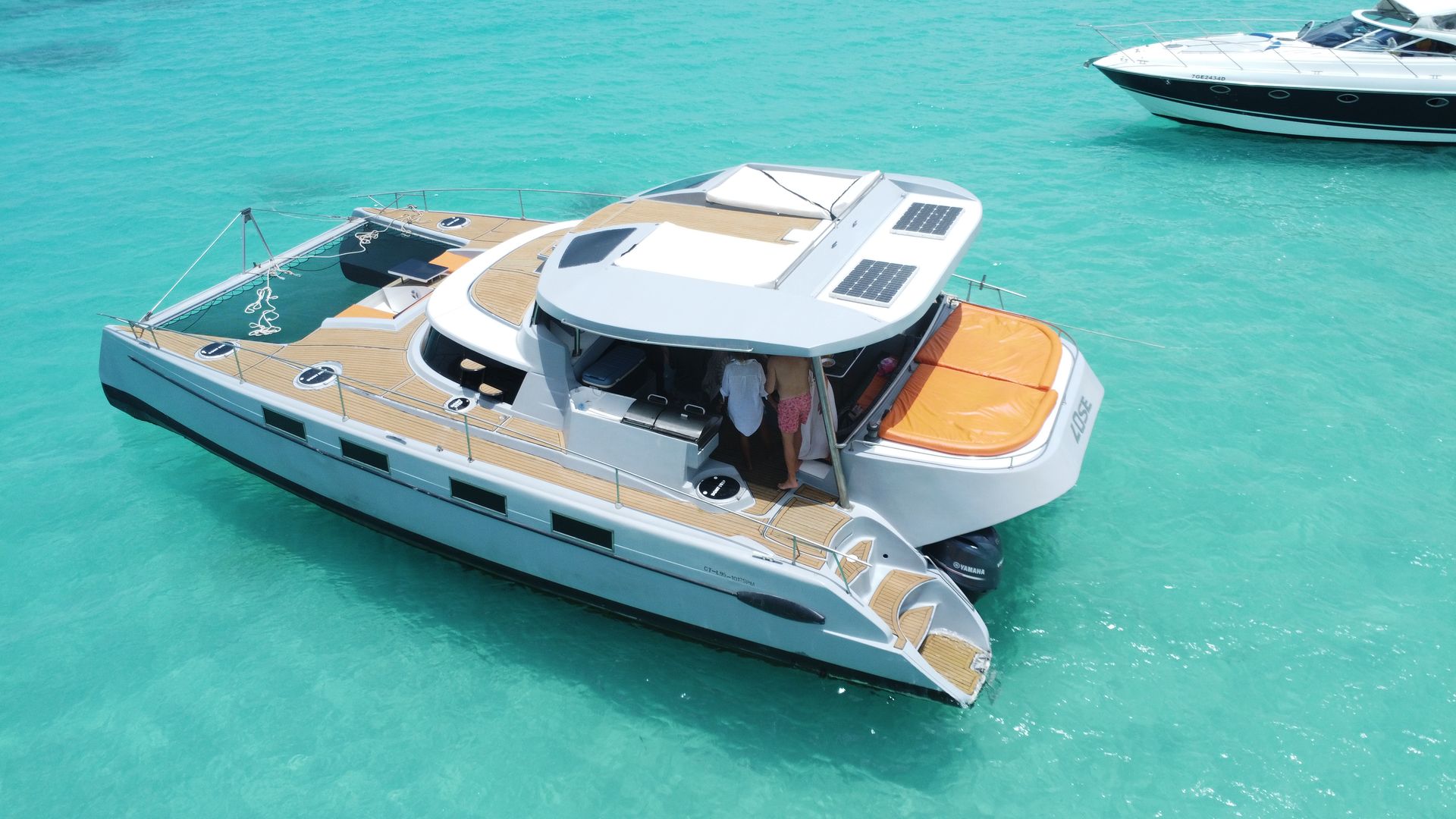 Aerial view of a white luxury catamaran yacht with teak decks and an orange sun pad, anchored in clear turquoise tropical water with two people in the shaded cockpit.