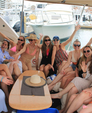 Group of friends in summer outfits and sunglasses cheering and waving aboard a yacht at a busy marina, straw sun hat on the table and other boats and docks visible in the background.