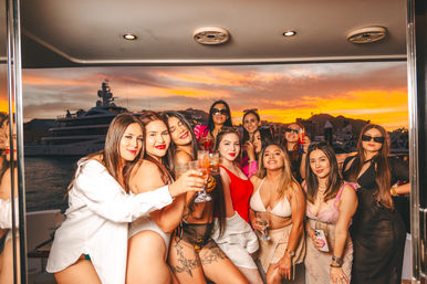 Group of women enjoying a sunset yacht party in a marina, raising drinks with luxury yachts and a golden coastal skyline in the background.