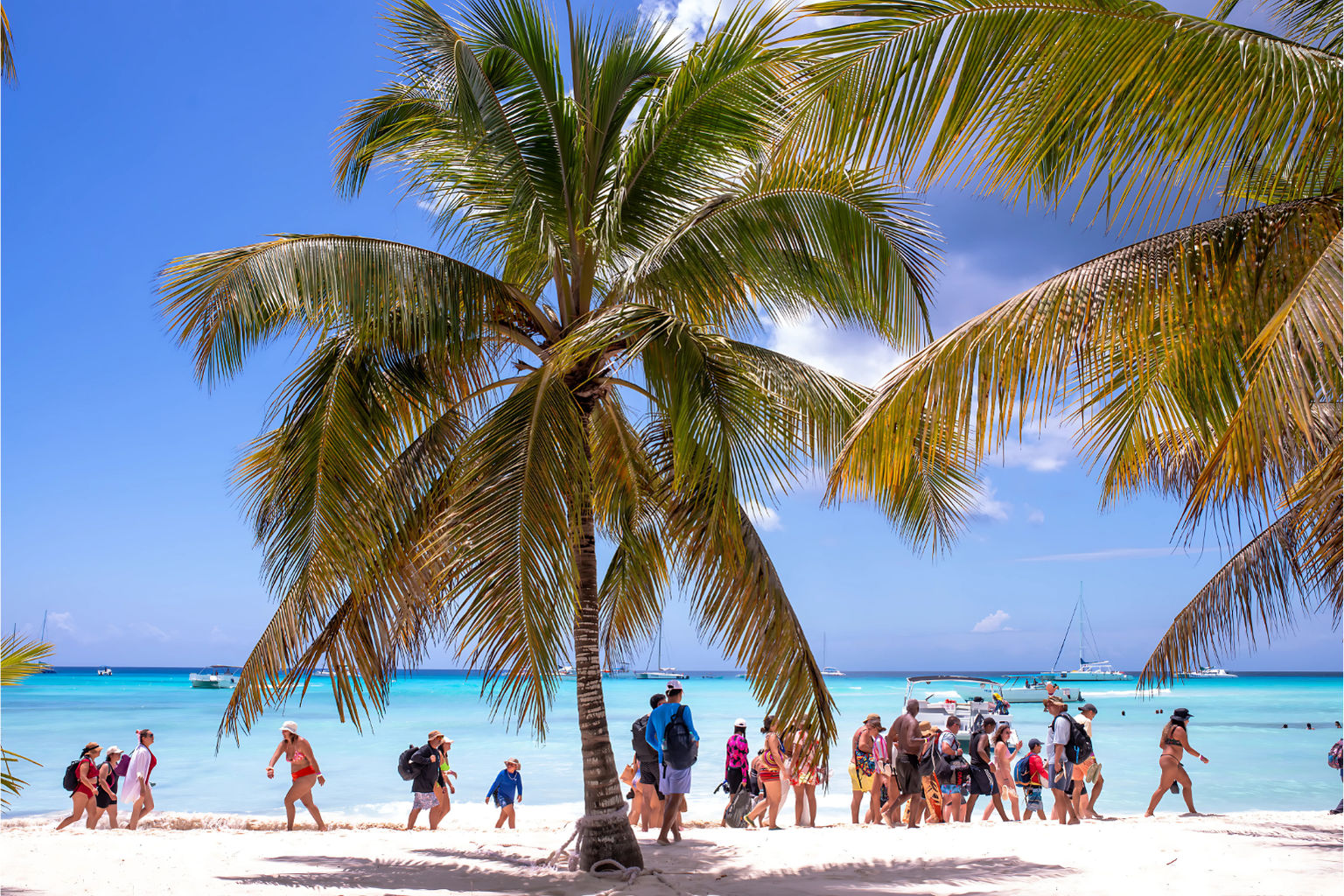 Sunny tropical beach with tall palm trees framing white sand and turquoise water, groups of beachgoers strolling along the shore and boats and sailboats anchored offshore under a bright blue sky.
