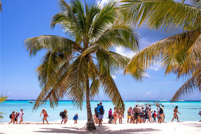 Sunny tropical beach with tall palm trees framing white sand and turquoise water, groups of beachgoers strolling along the shore and boats and sailboats anchored offshore under a bright blue sky.