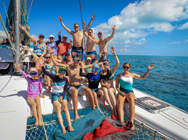 Cheerful multigenerational group of family and friends in swimsuits on a sunlit catamaran, waving and holding drinks against a bright blue sky and turquoise ocean backdrop.
