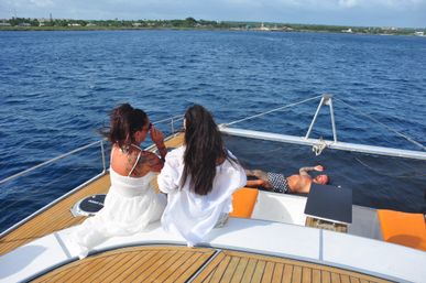 Two women in white outfits sitting on a yacht's teak deck looking out over deep blue ocean toward a distant green coastline, while a man in swim trunks relaxes on the bow netting under sunny skies.