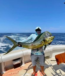Masked angler holding a large golden-green mahi-mahi (dolphinfish) on a sportfishing boat in the open ocean under a clear blue sky