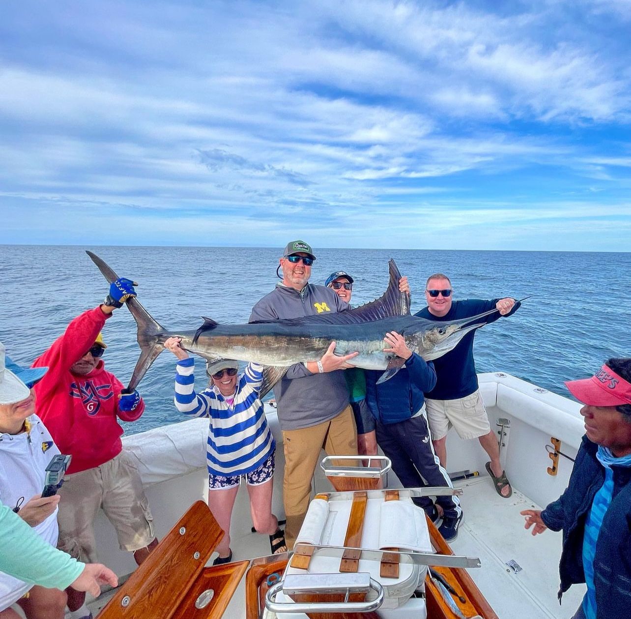 Group of anglers on a sportfishing boat holding a giant marlin (billfish) during offshore deep-sea fishing, calm blue ocean and partly cloudy sky on the horizon.