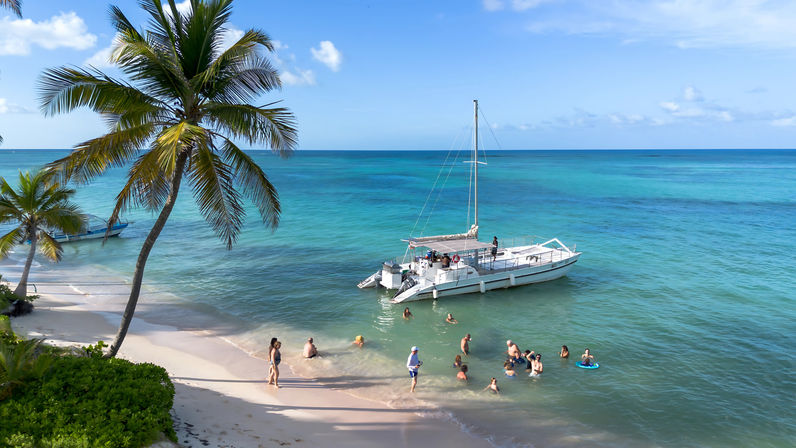 Tropical beach scene with leaning palm trees on white sand, turquoise sea and an anchored catamaran while people wade and swim under a bright blue sky