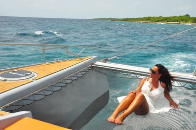 Woman in white dress lounging on a catamaran net above turquoise ocean with rocky green coastline in the distance