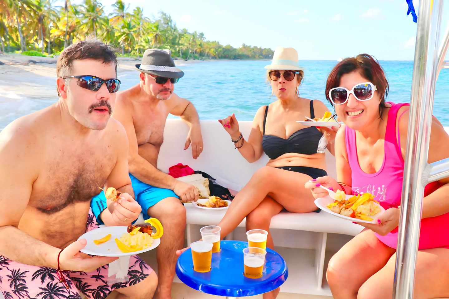Four adults enjoying a sunny boat picnic off a palm-lined tropical beach — wearing swimsuits and sunglasses, eating plates of grilled skewers and fries and sipping beers in plastic cups on turquoise water.