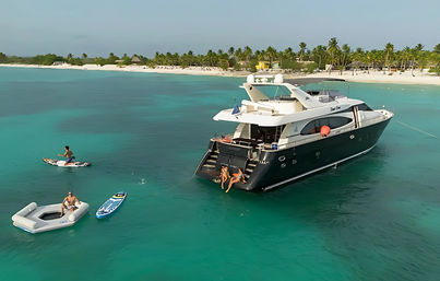 Luxury motor yacht anchored in turquoise lagoon off a palm‑fringed white‑sand beach, with people on the swim platform and using a paddleboard, inflatable and jetboard.