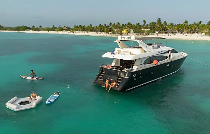 Luxury motor yacht anchored in turquoise lagoon off a palm‑fringed white‑sand beach, with people on the swim platform and using a paddleboard, inflatable and jetboard.