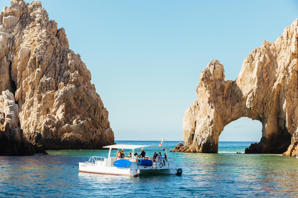 Sunlit tour boat gliding on turquoise water beside a dramatic natural rock arch and rugged sandstone cliffs under a clear blue sky