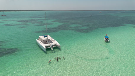 White catamaran anchored in shallow turquoise tropical lagoon with people swimming nearby and a small motorboat in the distance on a sunny day
