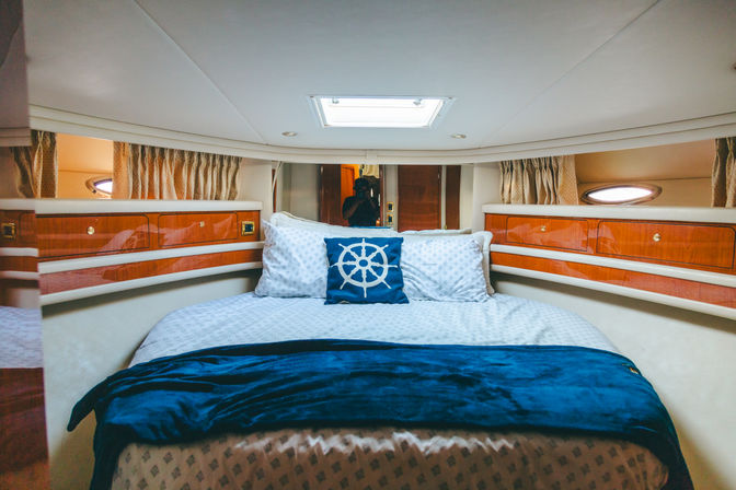 Cozy yacht cabin bedroom with a queen bed, nautical ship-wheel accent pillow, white patterned linens, navy throw blanket, glossy wood storage cabinets and oval porthole windows.