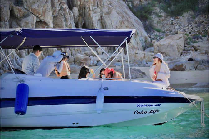 Group of friends on a white motorboat in turquoise water by a rocky coastal cove, sunlit beach outing with swimsuits and a photographer snapping photos.
