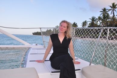 Smiling woman in a black dress seated on a catamaran deck with turquoise water and a palm-fringed tropical island in the background.