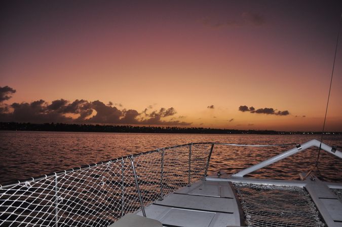 View from a catamaran bow at a golden-pink sunset over calm ocean, silhouetted palm-lined coastline and scattered clouds on the horizon.