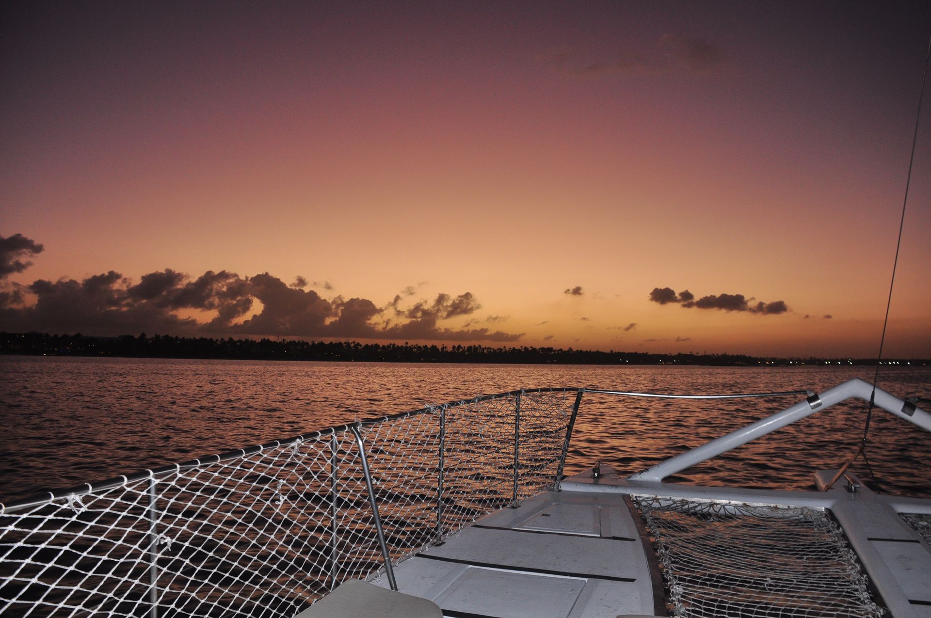 Purple-orange sunset over calm sea viewed from a catamaran bow, deck and safety netting in foreground with distant palm-lined shoreline silhouette.
