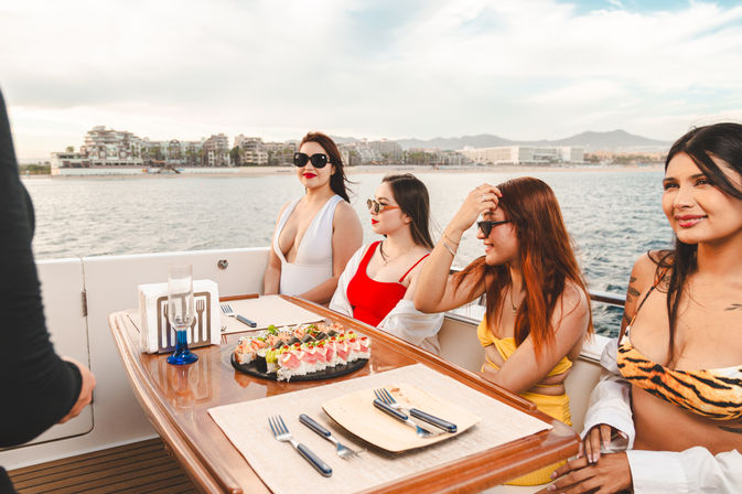Sunlit yacht brunch with four women in swimsuits and sunglasses enjoying a sushi platter and ocean views with a coastal resort skyline