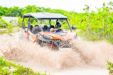 Orange off-road UTV tearing through a muddy puddle on a green trail, driver wearing a helmet and patterned face covering