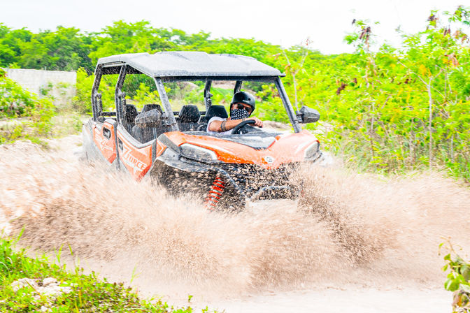Orange off-road UTV tearing through a muddy puddle on a green trail, driver wearing a helmet and patterned face covering