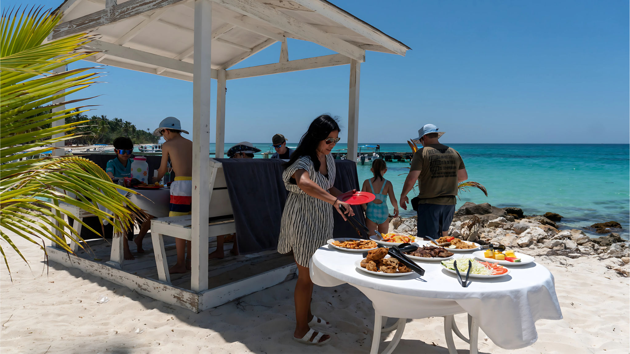 Woman serving from a beachside buffet under a weathered white cabana on a sunny tropical white-sand shore with turquoise water, palm fronds and families nearby enjoying the seaside.
