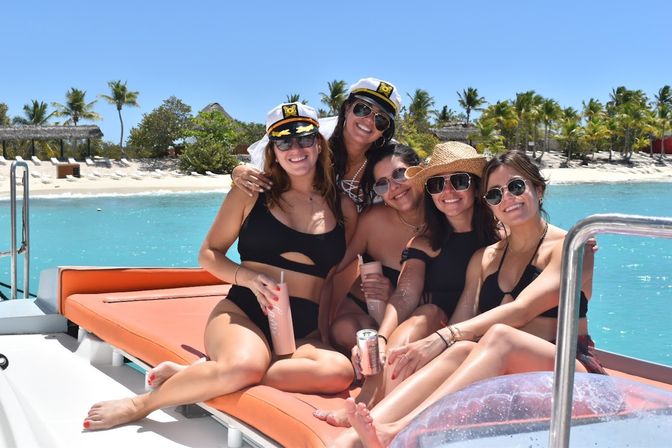 Five smiling friends in black swimsuits and sunglasses lounging on an orange sunbed aboard a boat, holding drinks and wearing captain and straw hats with turquoise water and a palm‑fringed white‑sand beach in the background.