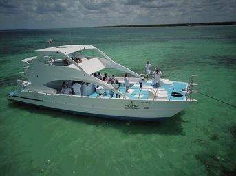 White luxury yacht with a group of people on deck in white outfits, anchored in clear turquoise tropical water near a low island under a partly cloudy sky